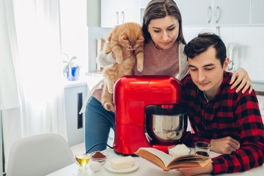 Young happy couple cooking on kitchen with food processor and holding cat. Woman and man reading recipe and hugging. Lifestyle