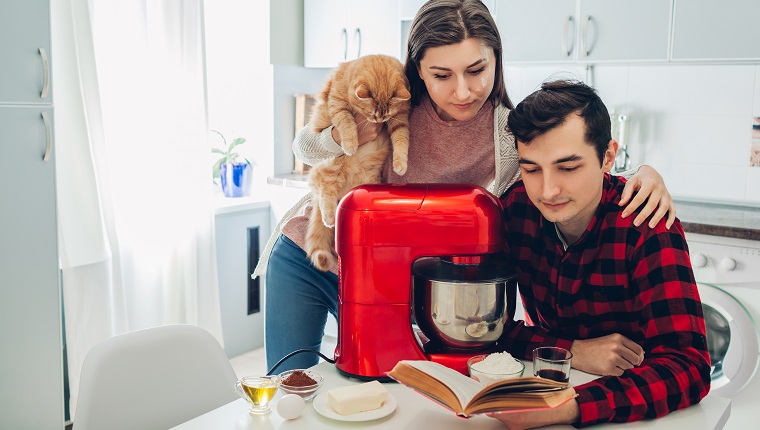 Young happy couple cooking on kitchen with food processor and holding cat. Woman and man reading recipe and hugging. Lifestyle