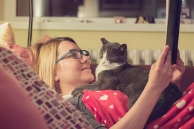 Beautiful young blonde woman is trying to read a book while her persistent cat is trying to sleep on her chest.