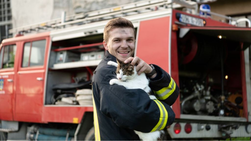 Close-up portrait of heroic fireman in protective suit and red helmet holds saved cat in his arms.